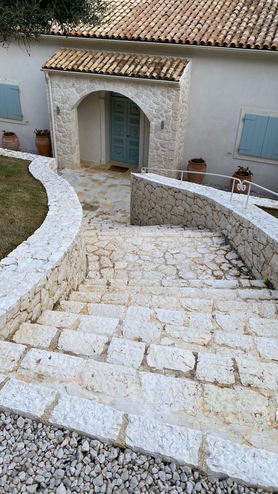 Curved cream stone steps and serpentine low walls descending from the arched entrance porch, pale blue-green shutters above.