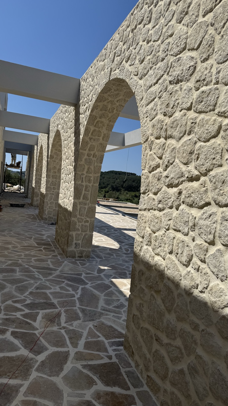 Looking down a colonnade of round fieldstone arches, white pergola beams overhead and flagstone paving below.